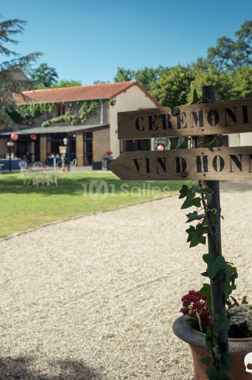 Panneau en bois indiquant ’Cérémonie’ et ’Vin d'honneur’ dans un jardin devant un bâtiment avec terrasse.
