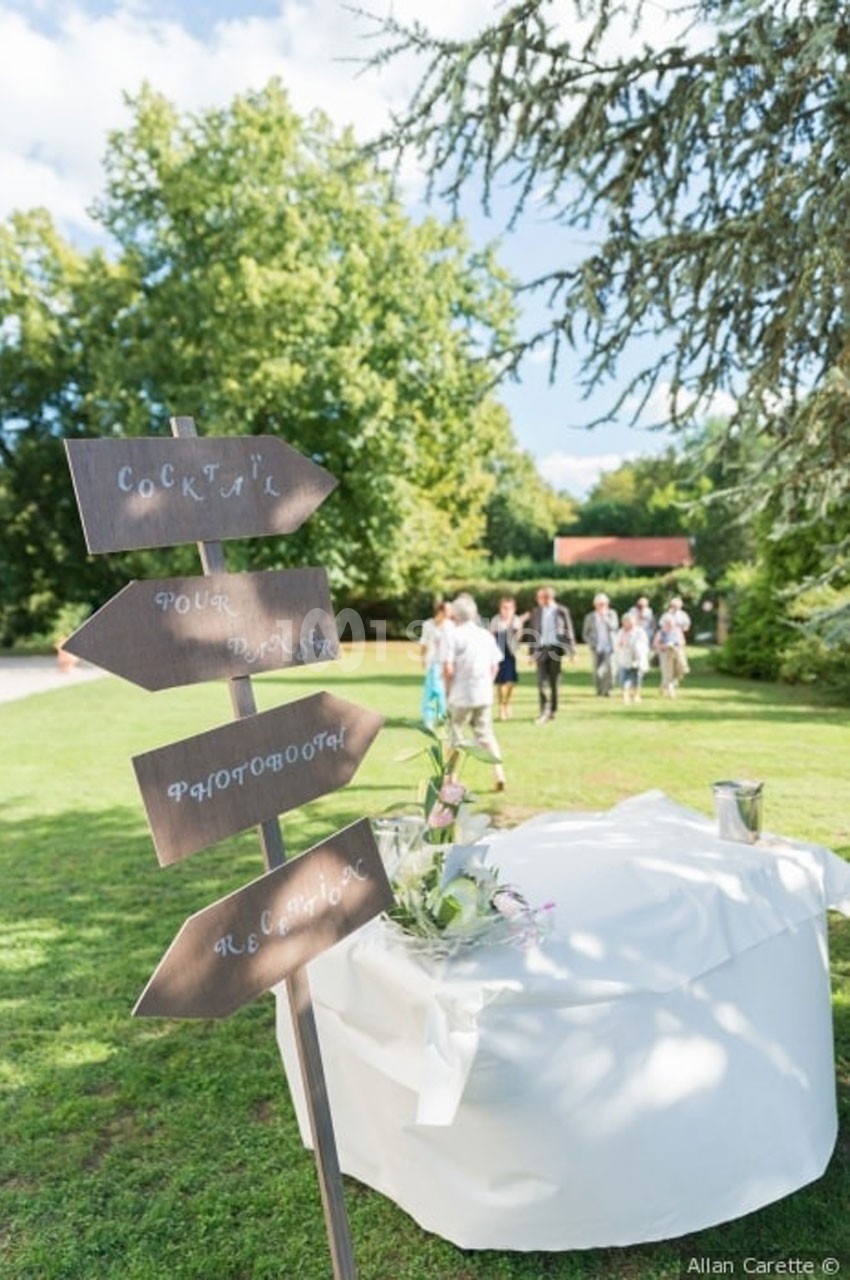 Panneaux directionnels en bois sur une pelouse, avec une table décorée et des invités en arrière-plan.