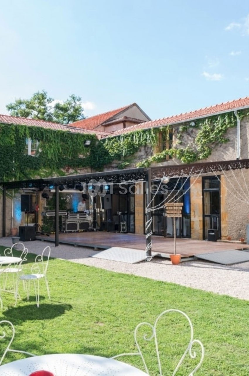 Cour extérieure avec pelouse, mobilier de jardin blanc, scène équipée pour un événement, bâtiment en pierre et lierre.