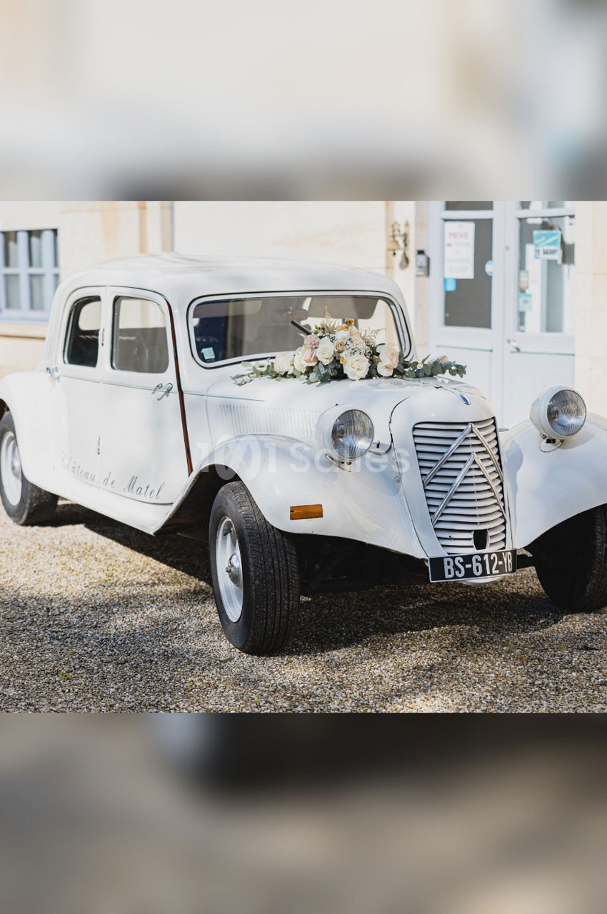 Voiture ancienne blanche décorée de fleurs blanches et roses, stationnée devant un bâtiment clair.