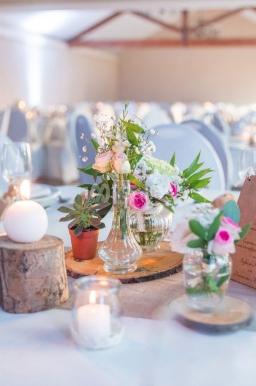 Décoration de table avec bouquets de fleurs, bougies et rondins de bois dans une salle de réception lumineuse.