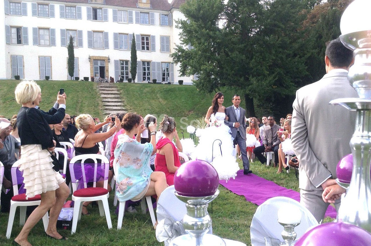 Un couple marche sur un tapis violet lors d'une cérémonie de mariage en plein air devant une grande maison.