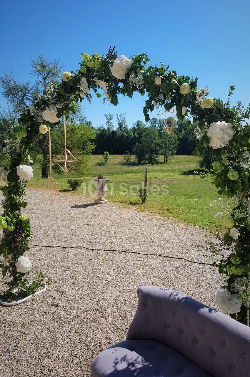Arche fleurie décorée de feuillage et de fleurs blanches, installée en extérieur sur un sol gravillonné.