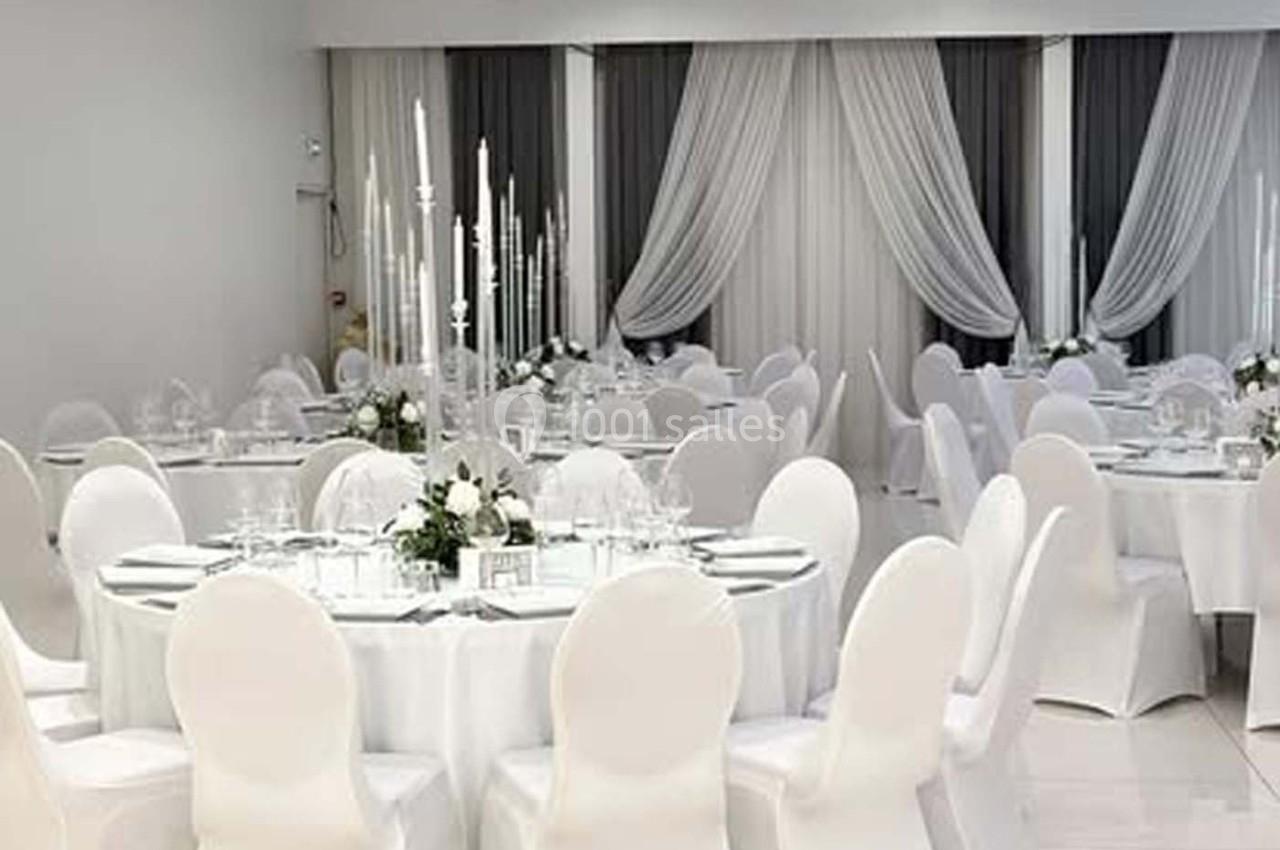 Salle de réception élégante avec tables rondes dressées, nappes blanches et chandeliers, décorée de rideaux clairs.