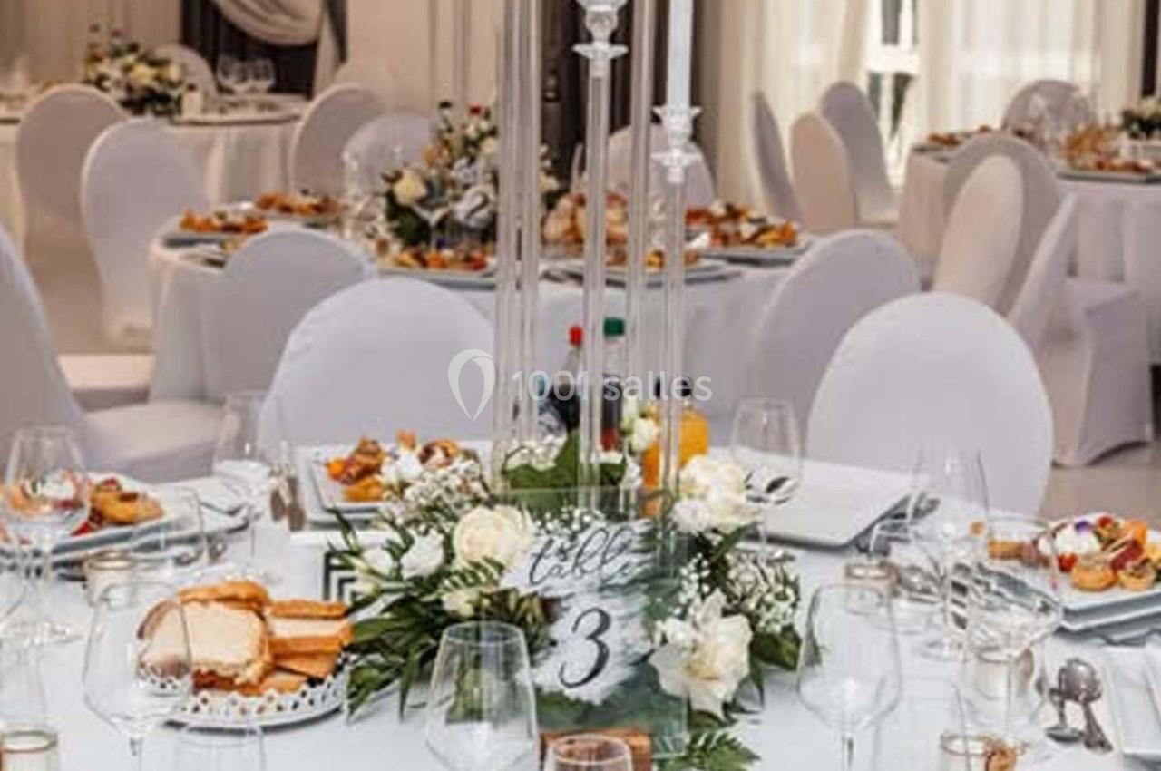 Salle de réception décorée avec des tables rondes, nappes blanches, centre de table floral et numéro de table 3.