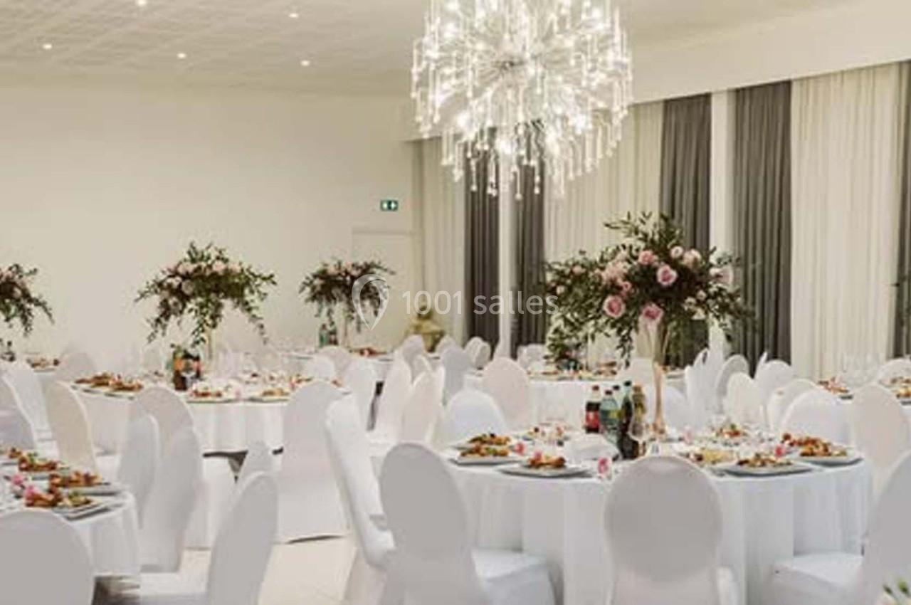 Salle de réception élégante avec tables rondes dressées, chaises blanches et décoration florale raffinée.