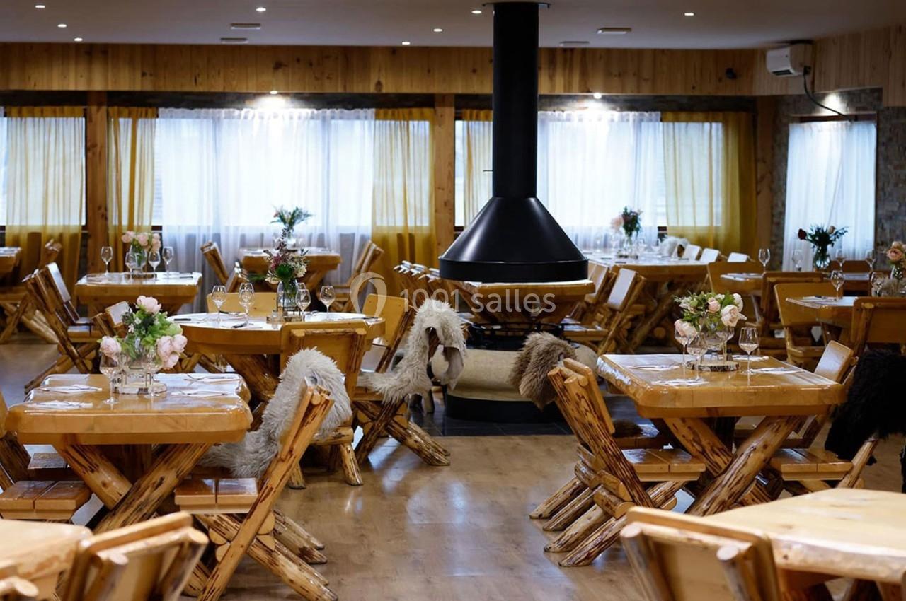 Salle de restaurant chaleureuse avec tables en bois, chaises décorées de fourrures et cheminée centrale noire.