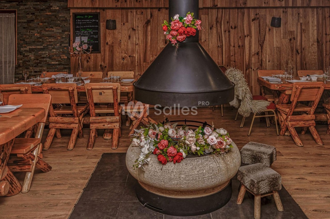Salle de restaurant rustique avec tables en bois, cheminée centrale décorée de fleurs et tabourets en fourrure.