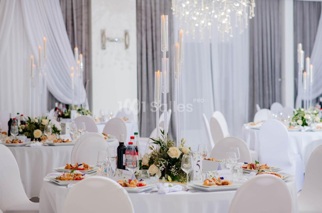 Salle de réception élégante avec tables rondes dressées, décorations florales et chandeliers modernes.