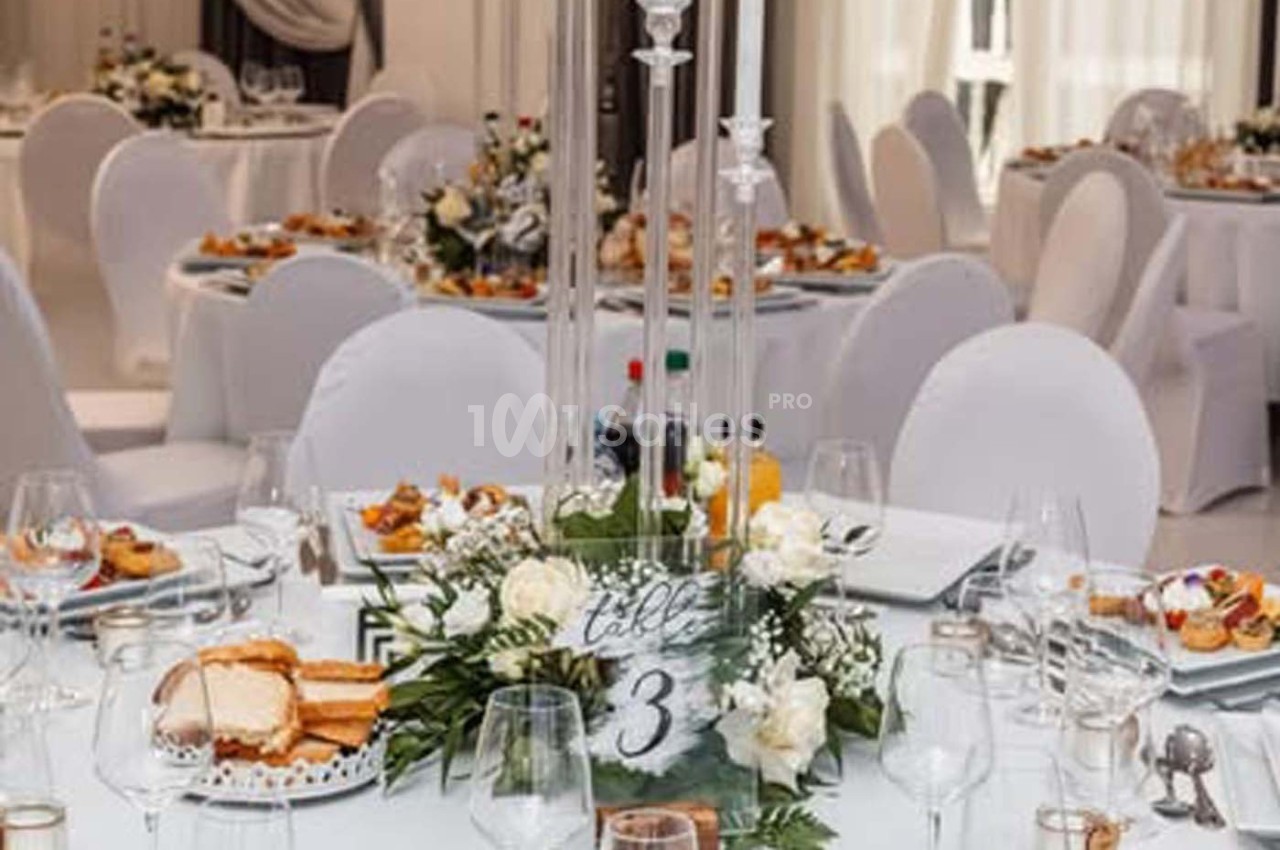 Salle de réception décorée avec des tables rondes, nappes blanches, centre de table floral et numéro de table 3.
