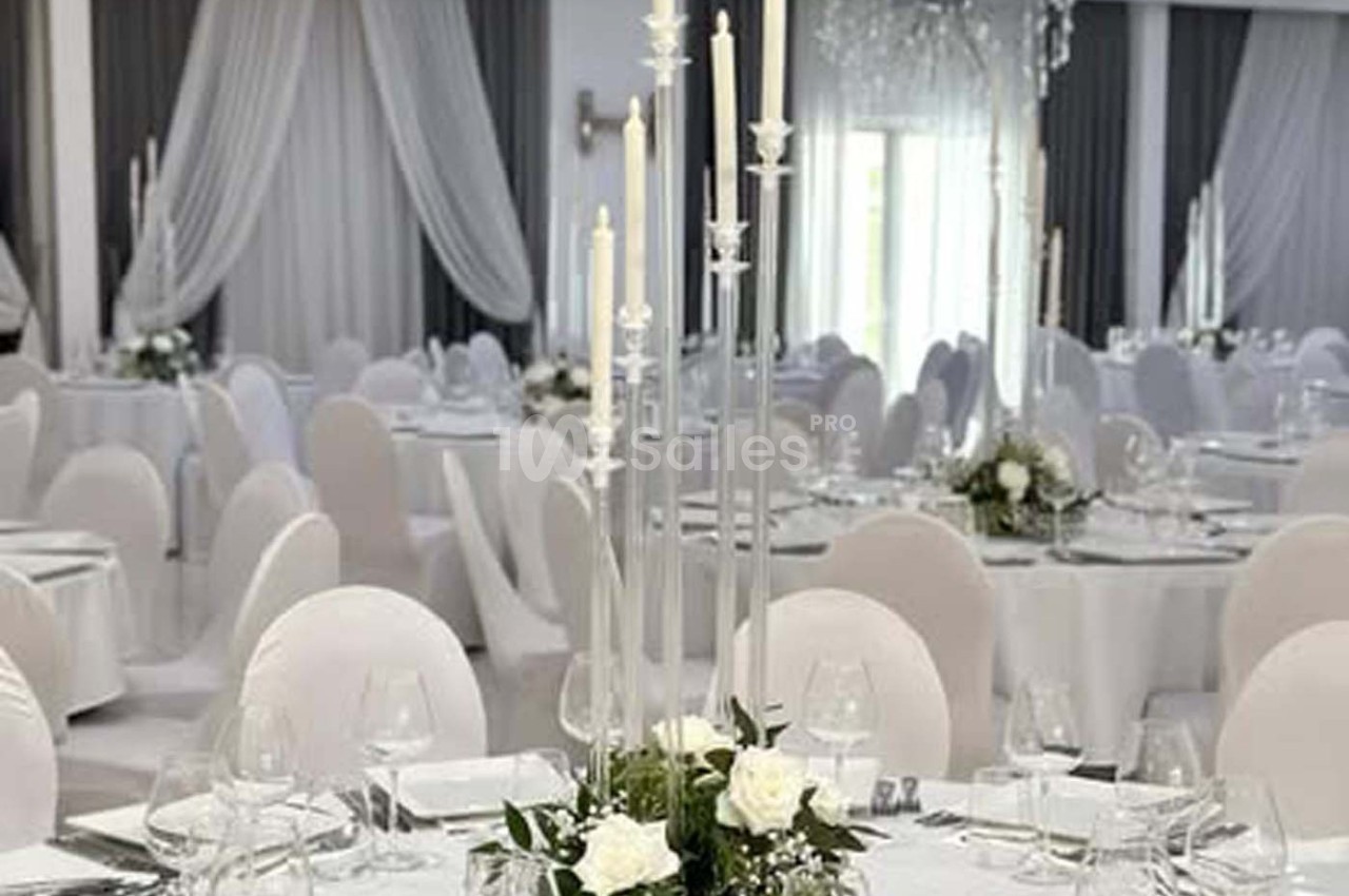 Salle de réception élégante avec tables rondes dressées, nappes blanches, chandeliers et décor floral.