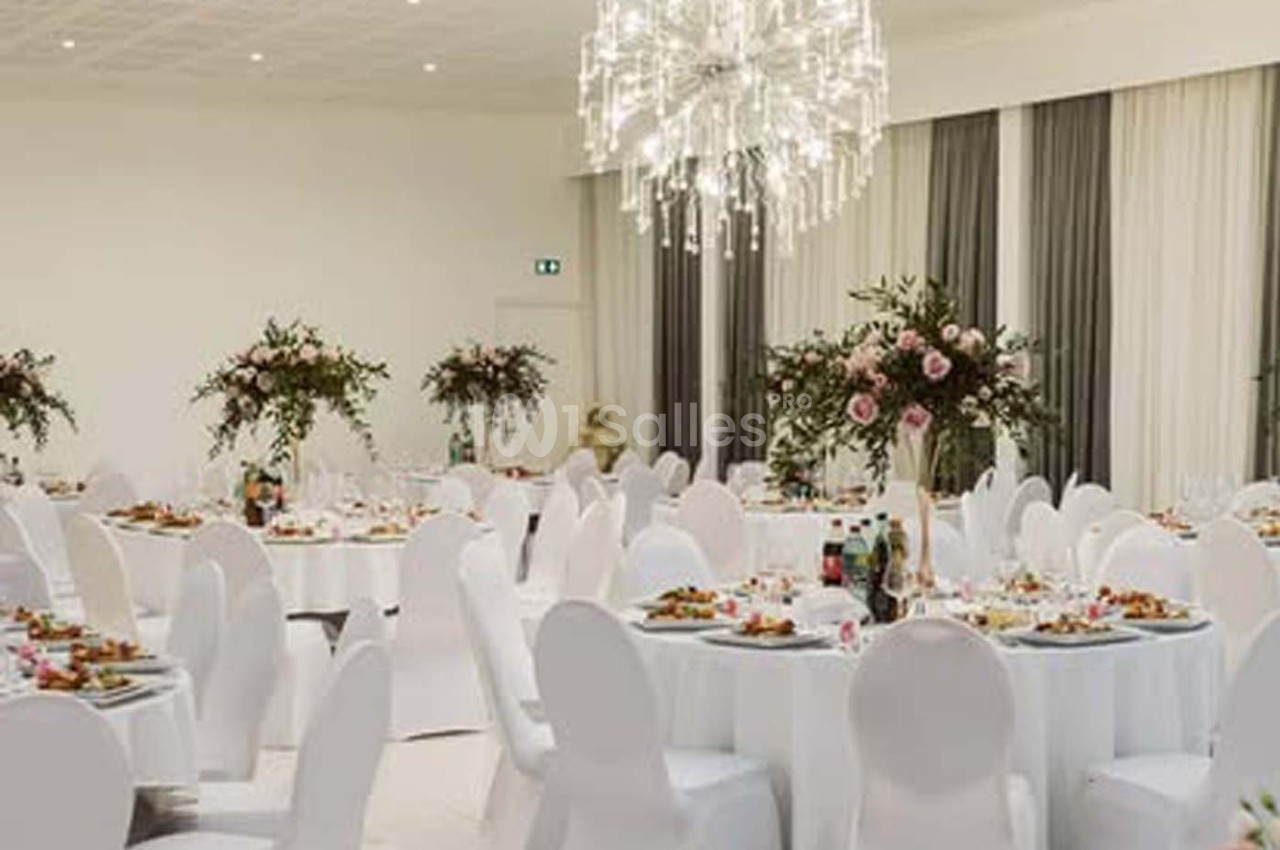Salle de réception élégante avec tables rondes dressées, chaises blanches et décorations florales.