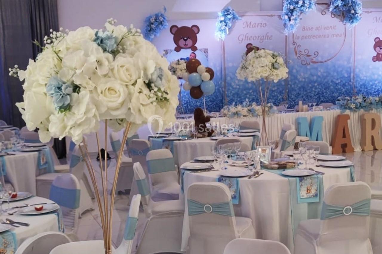 Salle décorée pour une fête avec des tables dressées, des fleurs blanches et bleues, et des éléments à thème ourson.