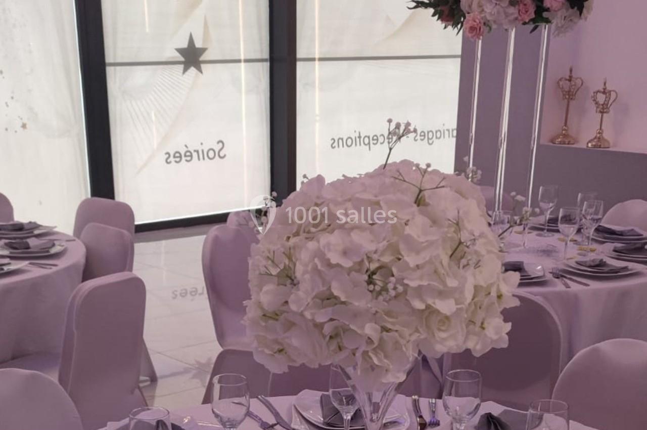 Salle de réception décorée avec des tables rondes, nappes blanches, centres de table fleuris et chaises assorties.
