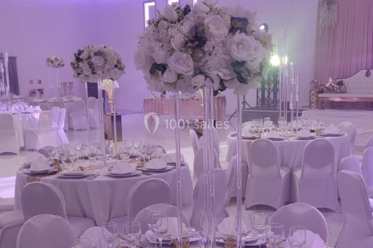 Salle de réception décorée avec des tables rondes, nappes blanches, centres de table fleuris et éclairage violet.