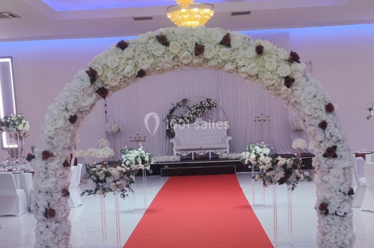 Arche florale blanche avec des roses rouges, tapis rouge menant à une scène décorée pour un événement festif.