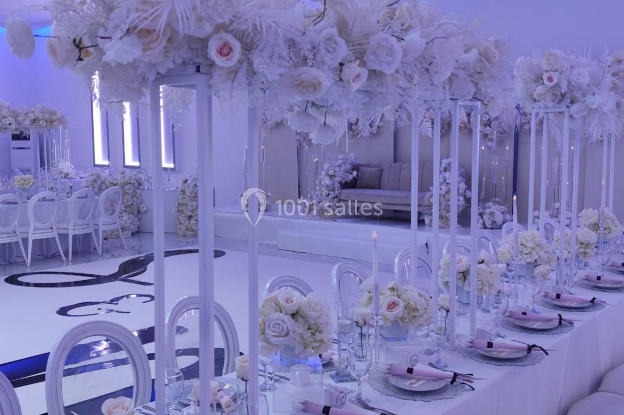 Salle de réception décorée avec des fleurs blanches, une longue table dressée et un éclairage bleu tamisé.
