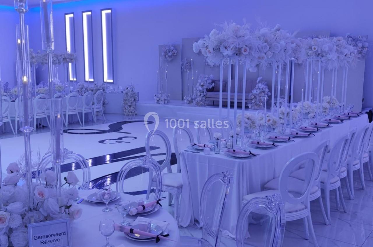 Salle de réception décorée en blanc avec des tables élégantes, des chaises transparentes et des arrangements floraux.