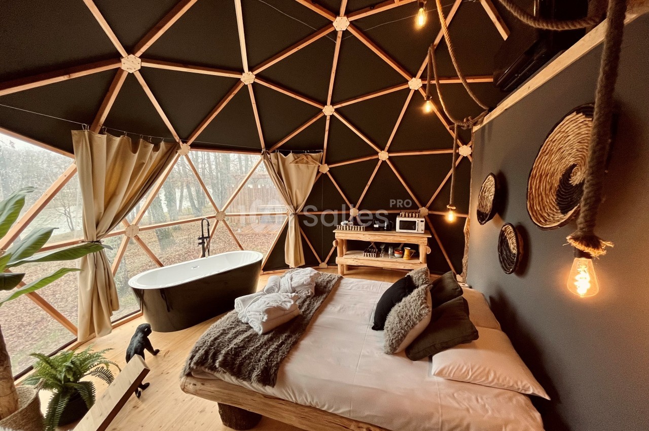 Intérieur d'un dôme géodésique avec lit en bois, baignoire, rideaux ouverts sur un paysage boisé et décoration naturelle.