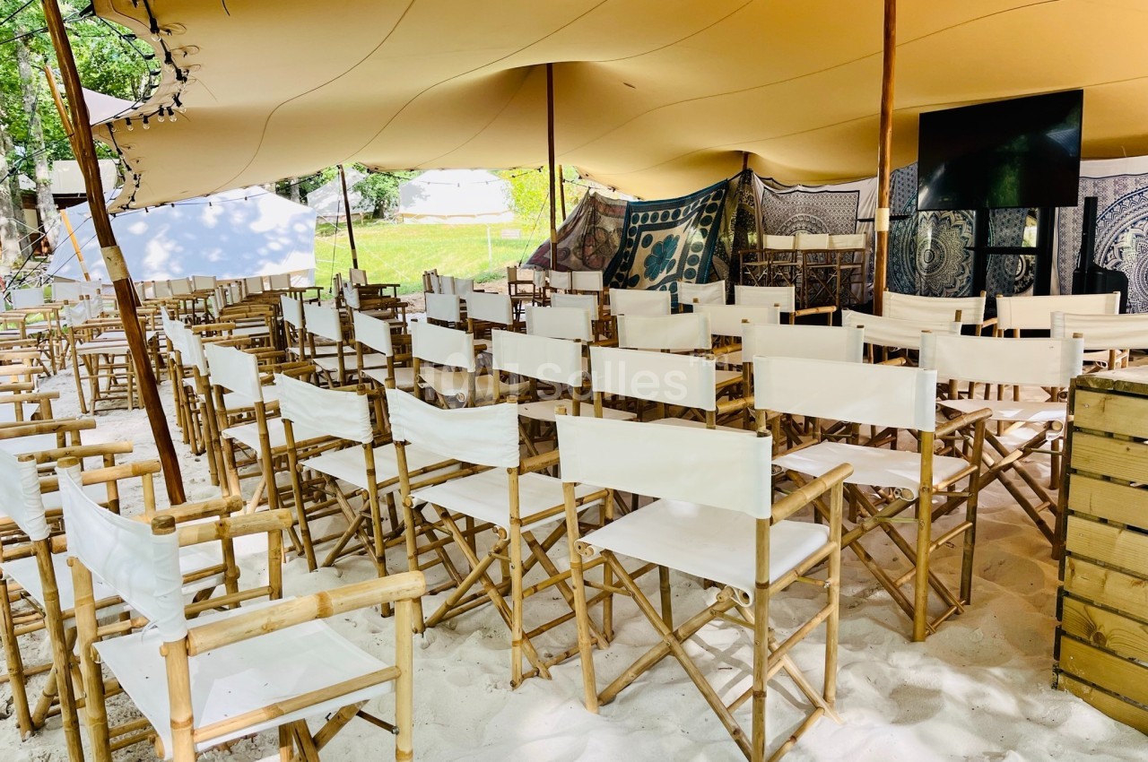 Chaises pliantes en bois avec assises blanches disposées sous une tente beige dans un espace extérieur sur du sable.