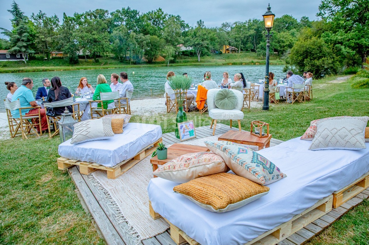 Des convives dînent en plein air près d'un lac, avec des lits de palettes décorés de coussins au premier plan.