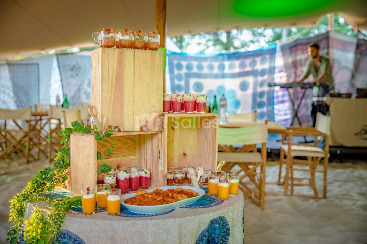 Buffet en plein air avec verrines colorées, plats variés et décor boisé, sur fond de tente et musicien en arrière-plan.