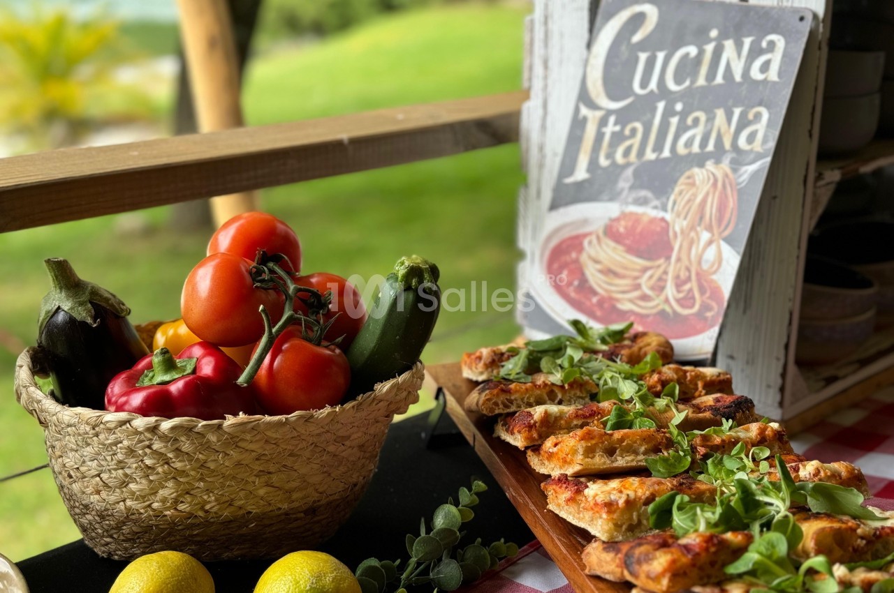 Panier de légumes frais à côté de mini-pizzas garnies, posé sur une table en plein air avec un panneau ’Cucina Italiana’.