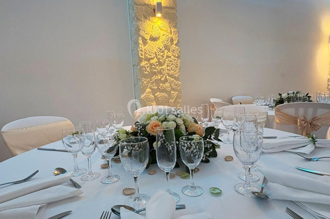 Table ronde élégamment dressée avec nappes blanches, verres, couverts et un centre de table floral dans une salle lumineuse.
