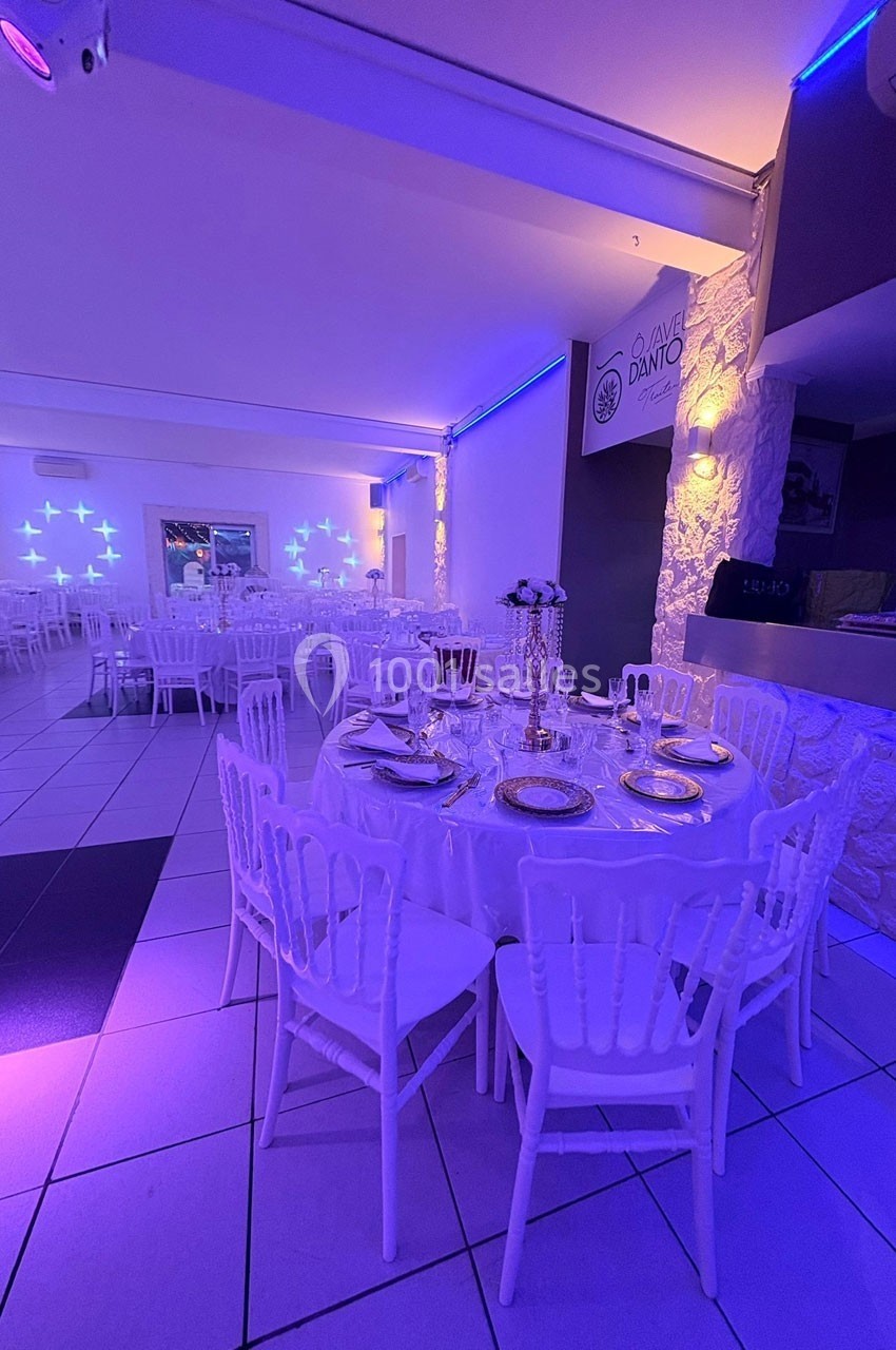 Salle de réception élégante avec tables rondes dressées, chaises blanches et éclairage tamisé violet.