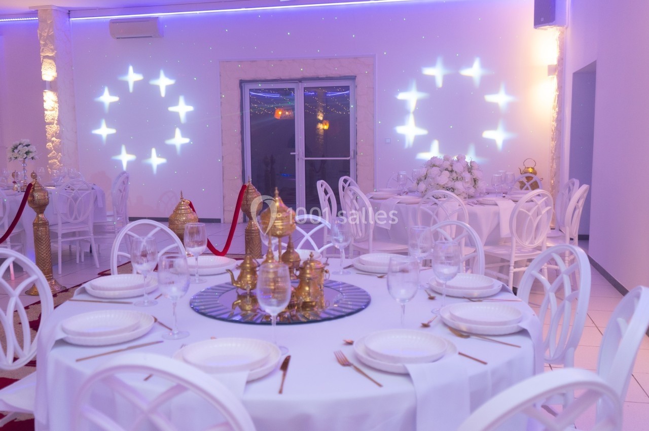 Salle de réception décorée avec des tables rondes dressées, des chaises blanches et des lumières d'ambiance.