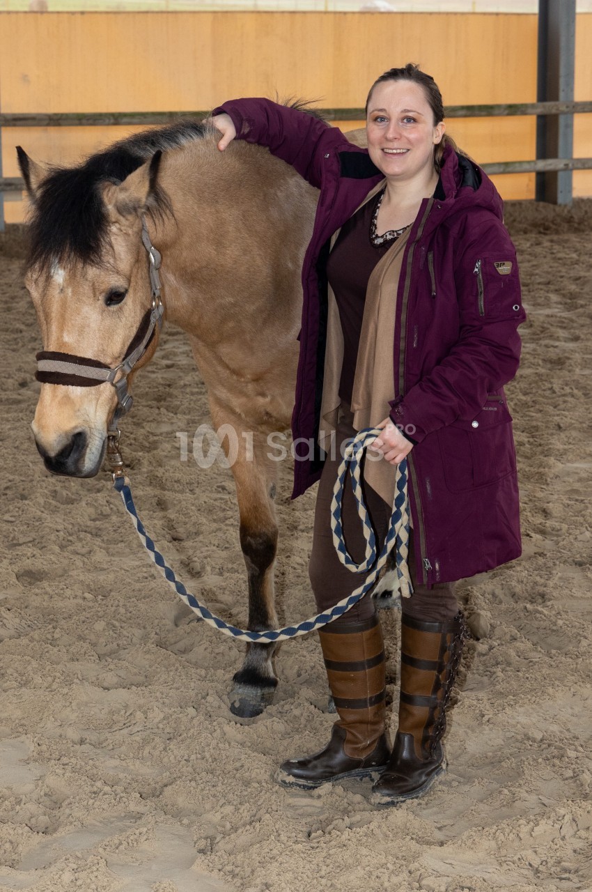 Une femme souriante tient un cheval par une longe dans un manège intérieur sur un sol sablonneux.