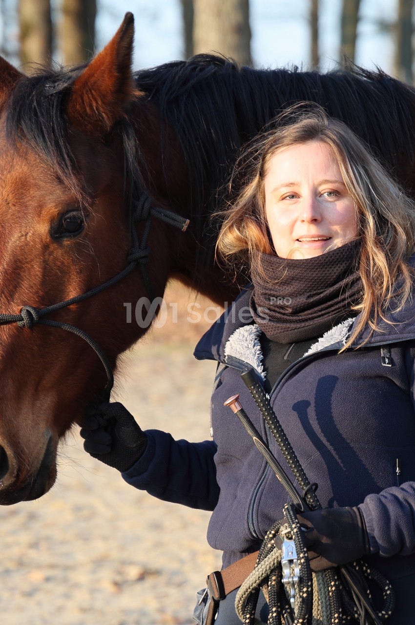 Femme souriante tenant les rênes d'un cheval brun dans un environnement extérieur boisé.