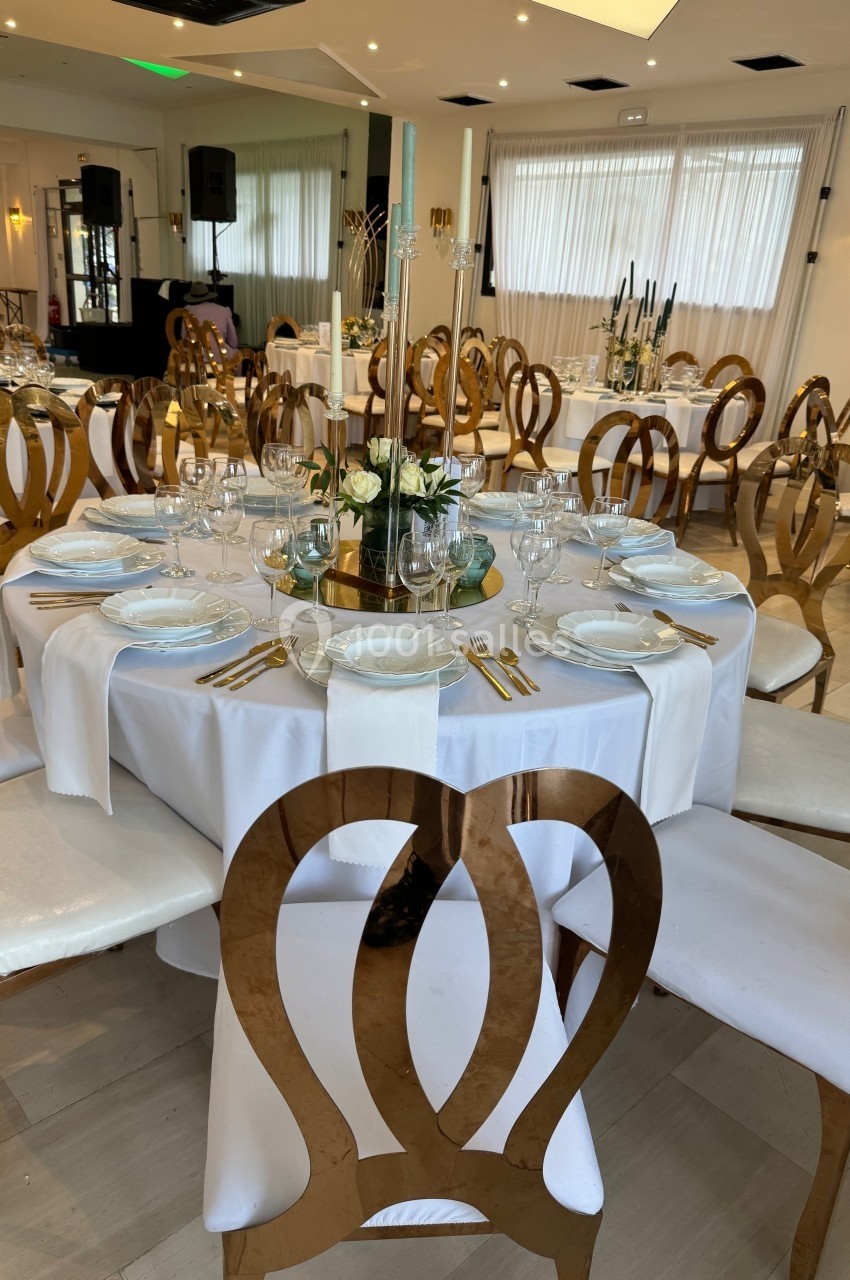 Salle de réception avec tables rondes dressées, nappes blanches, chaises dorées et décoration florale centrale.
