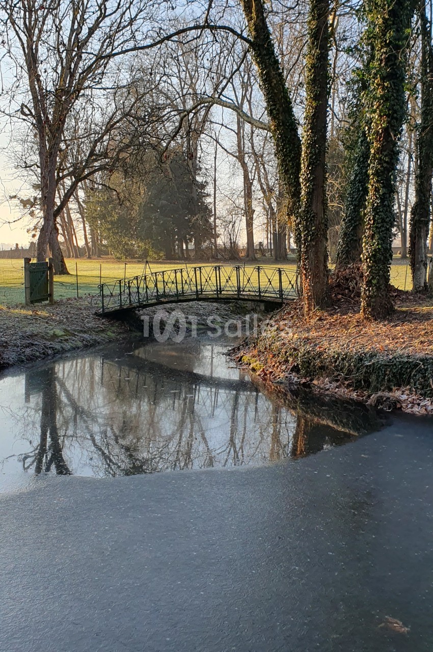 Petit pont en métal traversant un ruisseau entouré d'arbres en hiver, avec un champ en arrière-plan.