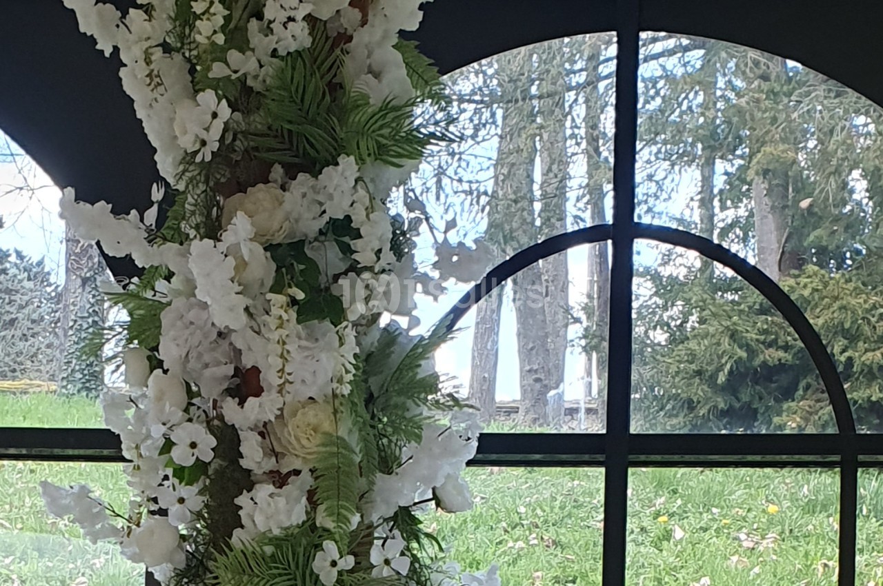 Arrangement floral blanc et vert devant une fenêtre cintrée donnant sur un jardin avec des arbres.