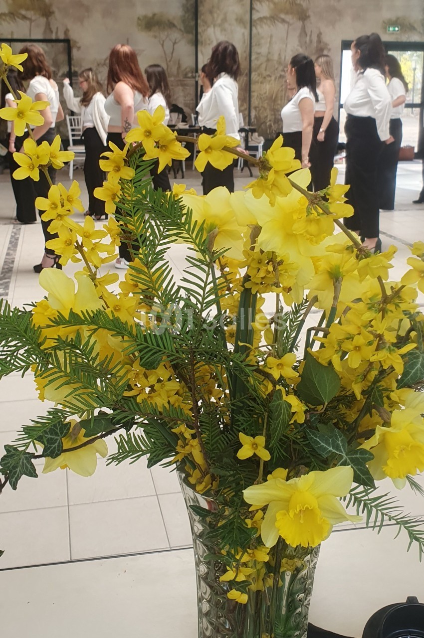 Un bouquet de fleurs jaunes dans un vase en verre, avec des personnes debout en arrière-plan dans une salle lumineuse.