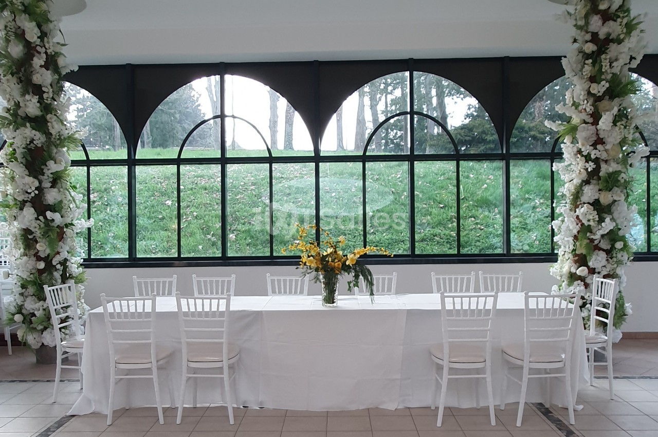 Salle lumineuse avec une grande table blanche décorée de fleurs jaunes, entourée de chaises blanches et de colonnes fleuries.
