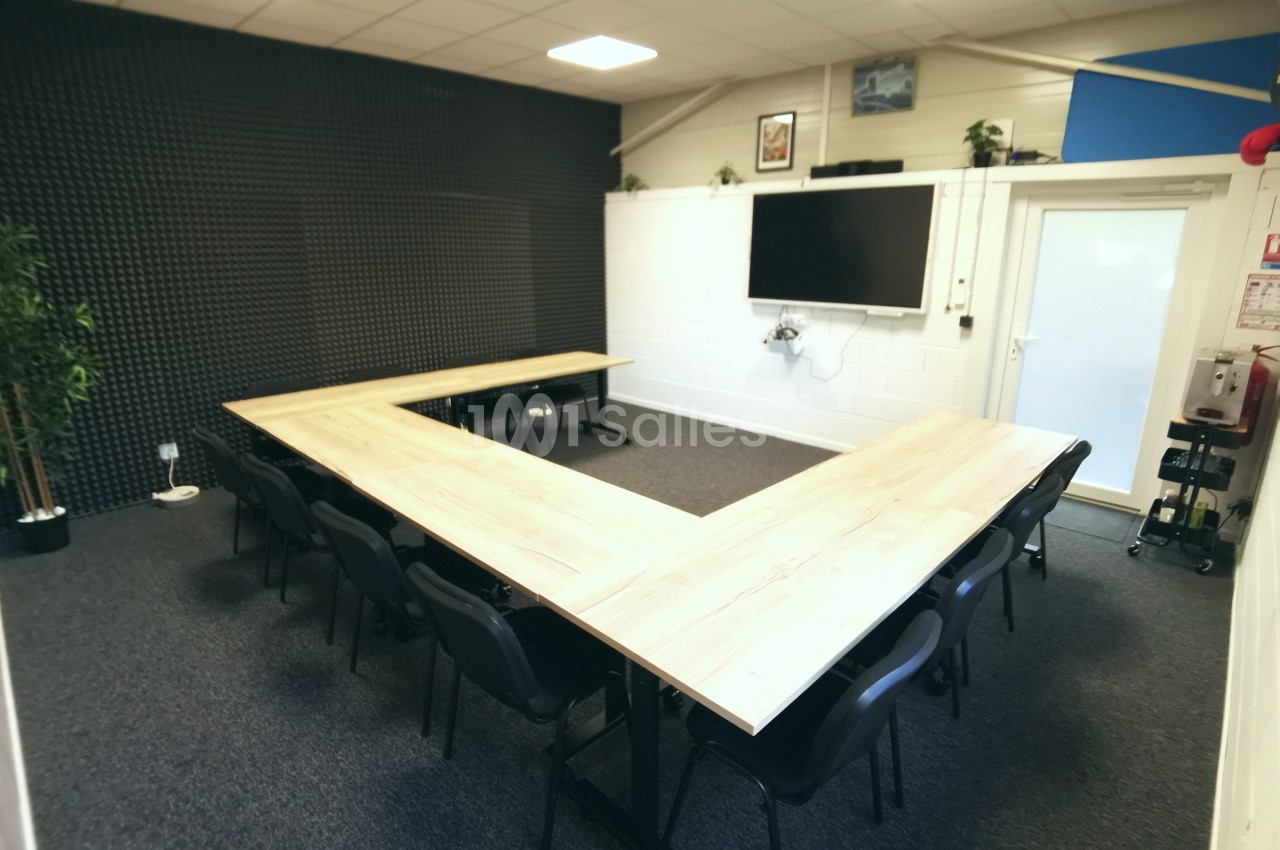 Salle de réunion avec tables en forme de U, chaises noires, écran mural et éclairage au plafond.