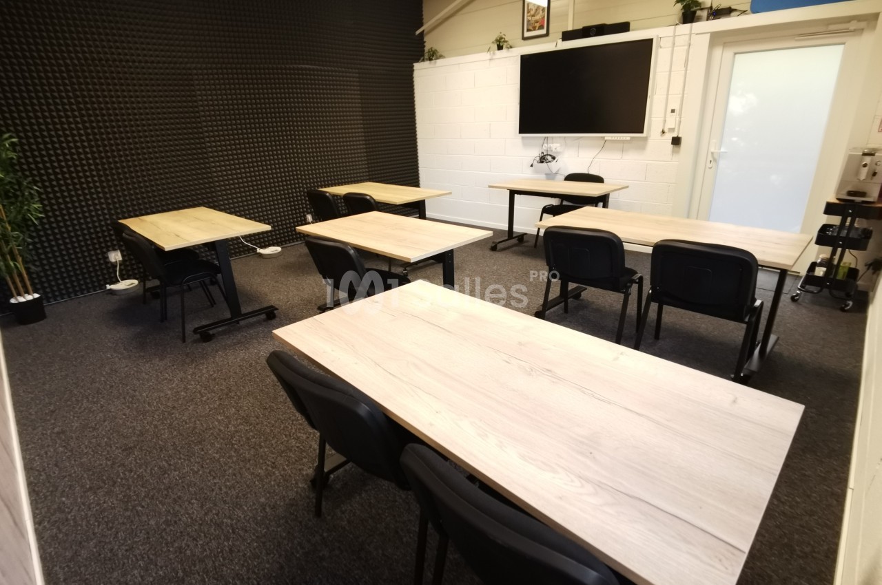 Salle de réunion équipée de tables en bois clair, chaises noires, écran mural et murs insonorisés.