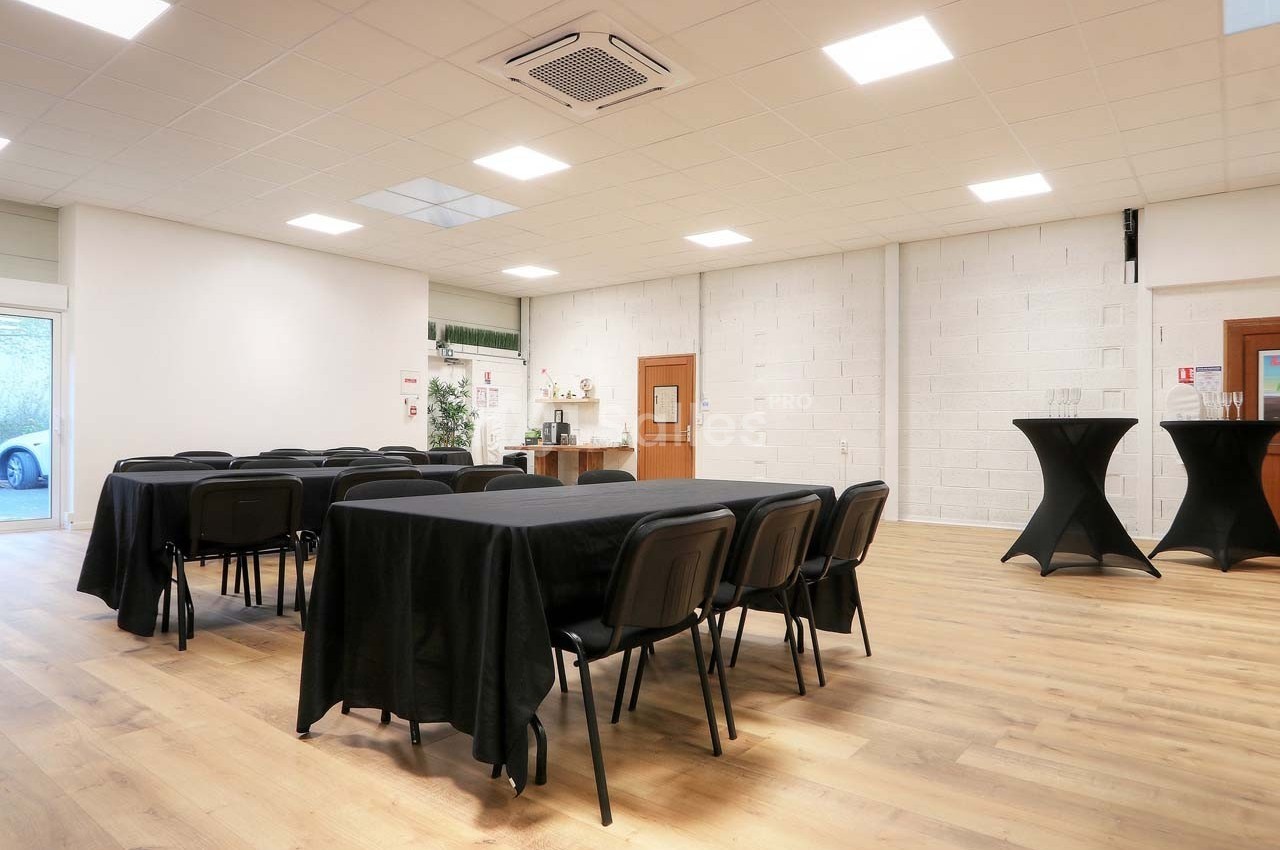 Salle lumineuse avec tables et chaises noires, sol en parquet, murs blancs et coin avec tables hautes.