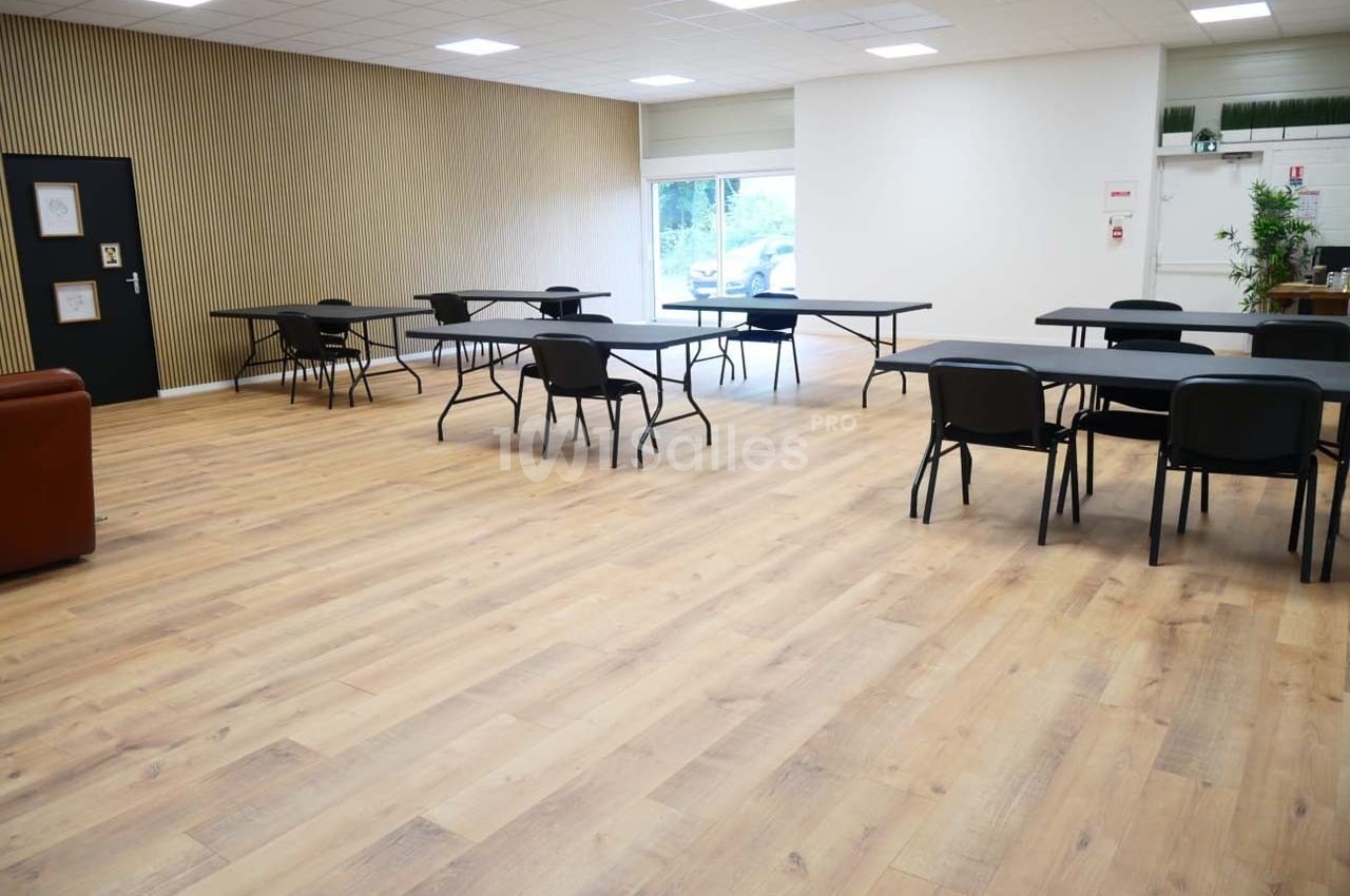 Salle lumineuse avec parquet, tables et chaises espacées, murs clairs et une baie vitrée donnant sur l'extérieur.