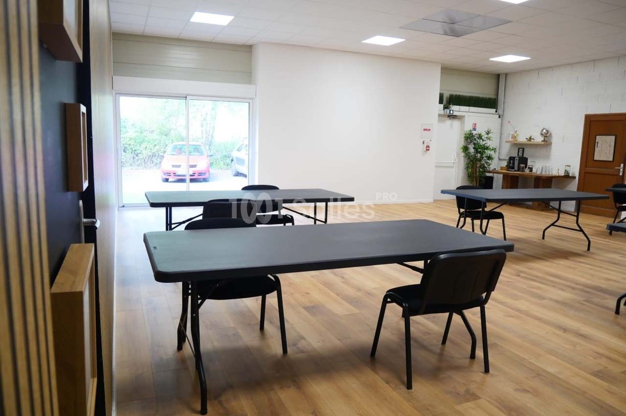 Salle lumineuse avec tables noires espacées, chaises, sol en bois, et une baie vitrée donnant sur l'extérieur.