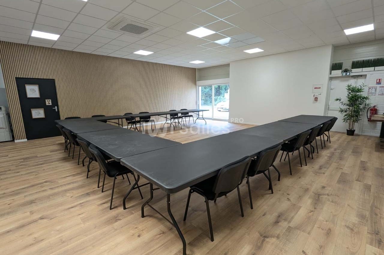 Salle de réunion lumineuse avec tables disposées en U, chaises noires et sol en parquet clair.
