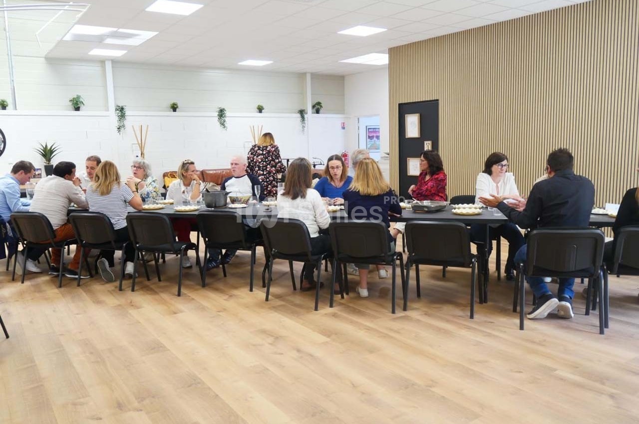 Des personnes assises autour de longues tables dans une salle lumineuse, partageant un repas ou une discussion.