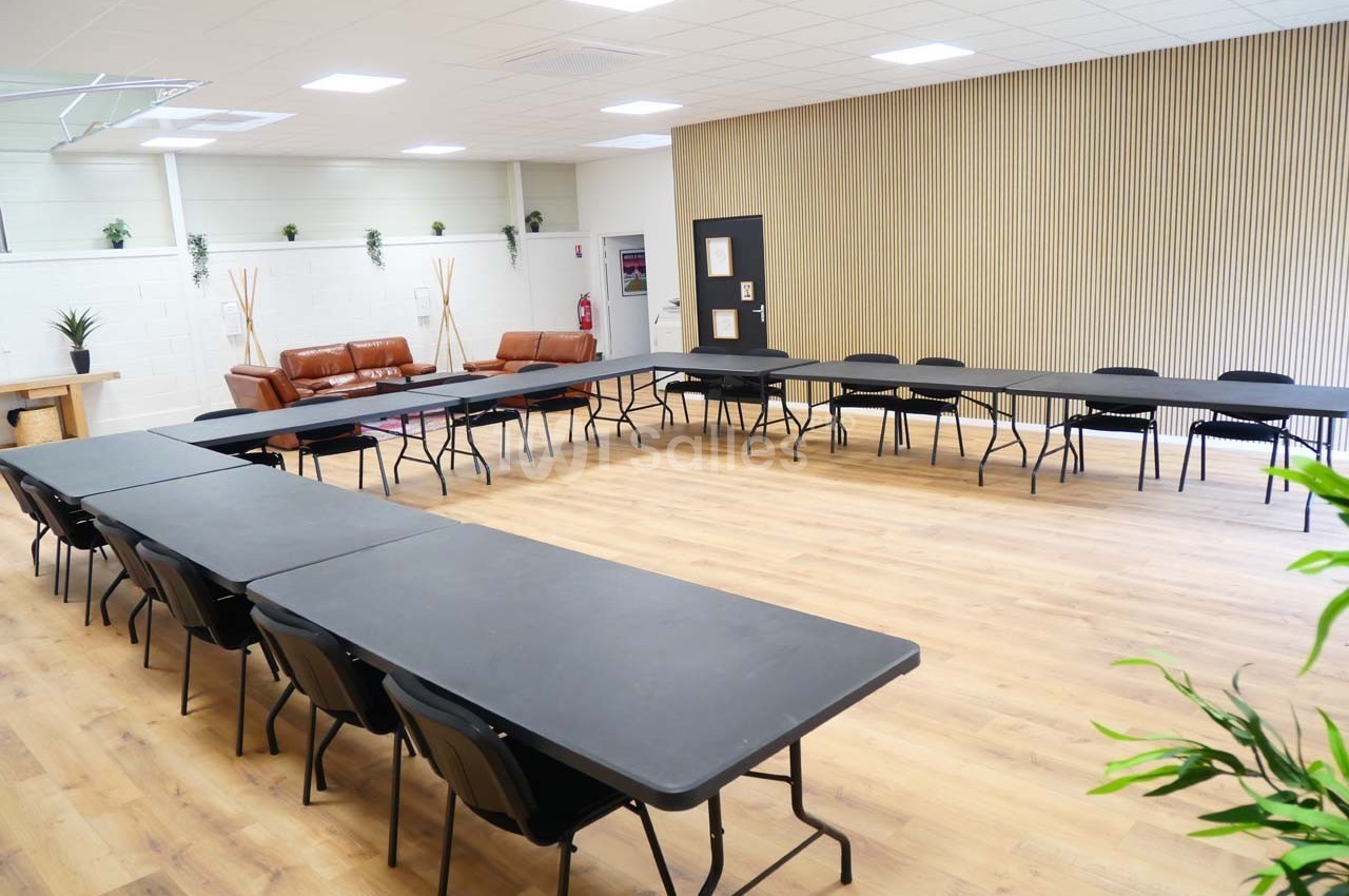 Salle lumineuse avec tables noires disposées en U, chaises alignées, parquet clair et mur décoré de lattes en bois.