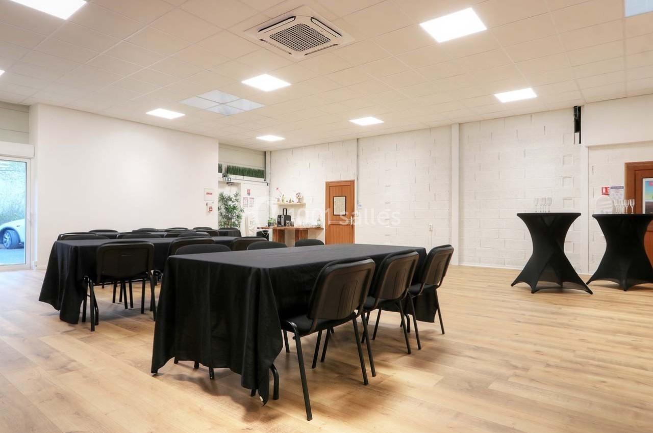 Salle lumineuse avec tables et chaises noires, sol en parquet, murs blancs et coin avec tables hautes.