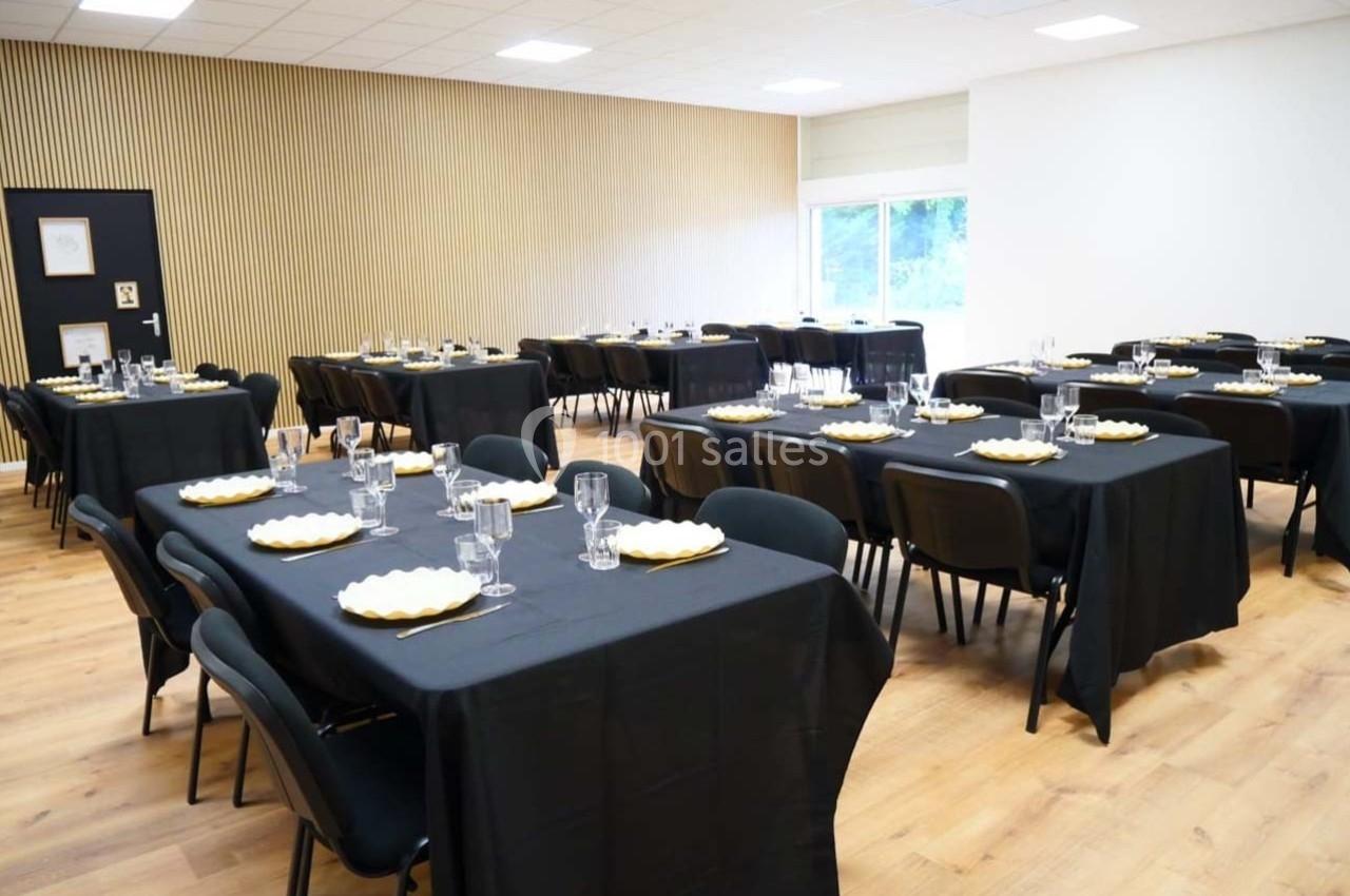 Salle aménagée avec des tables noires dressées, assiettes et verres, éclairée par une grande fenêtre.
