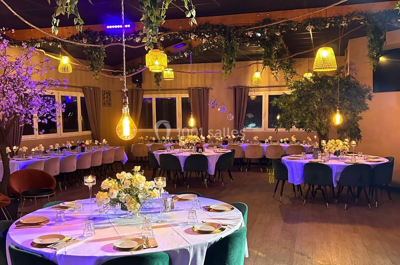 Salle de réception décorée avec des tables rondes dressées, éclairage chaleureux et végétation suspendue.