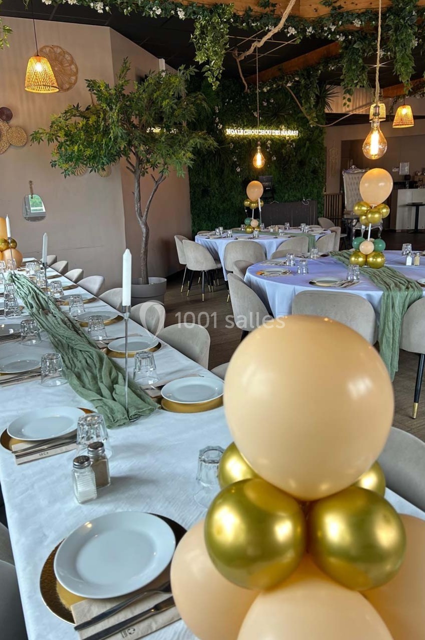 Salle décorée pour un événement avec tables dressées, nappes blanches, ballons dorés et beige, et végétation suspendue.