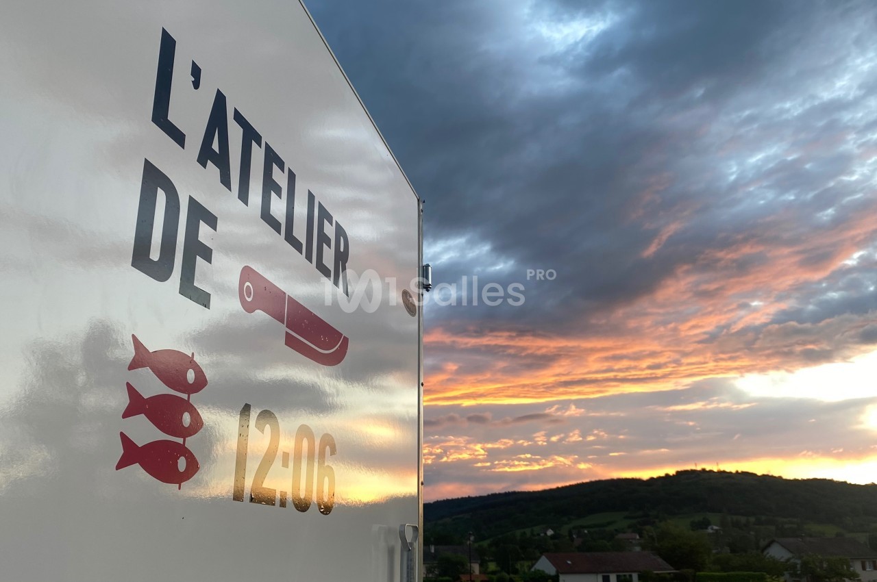 Flanc d'un camion marqué ’L'Atelier de 12:06’ avec un couteau et des poissons, devant un coucher de soleil champêtre.